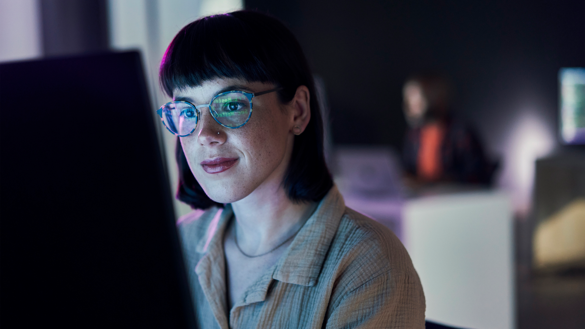 A woman with glasses smiles slightly while working at a computer in a blue-lit modern office.