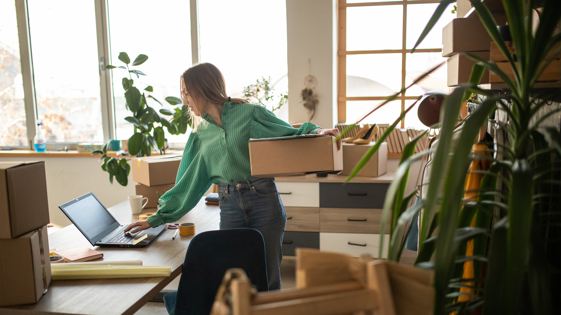 A person holding a box in an office with packing materials and a laptop on the desk.