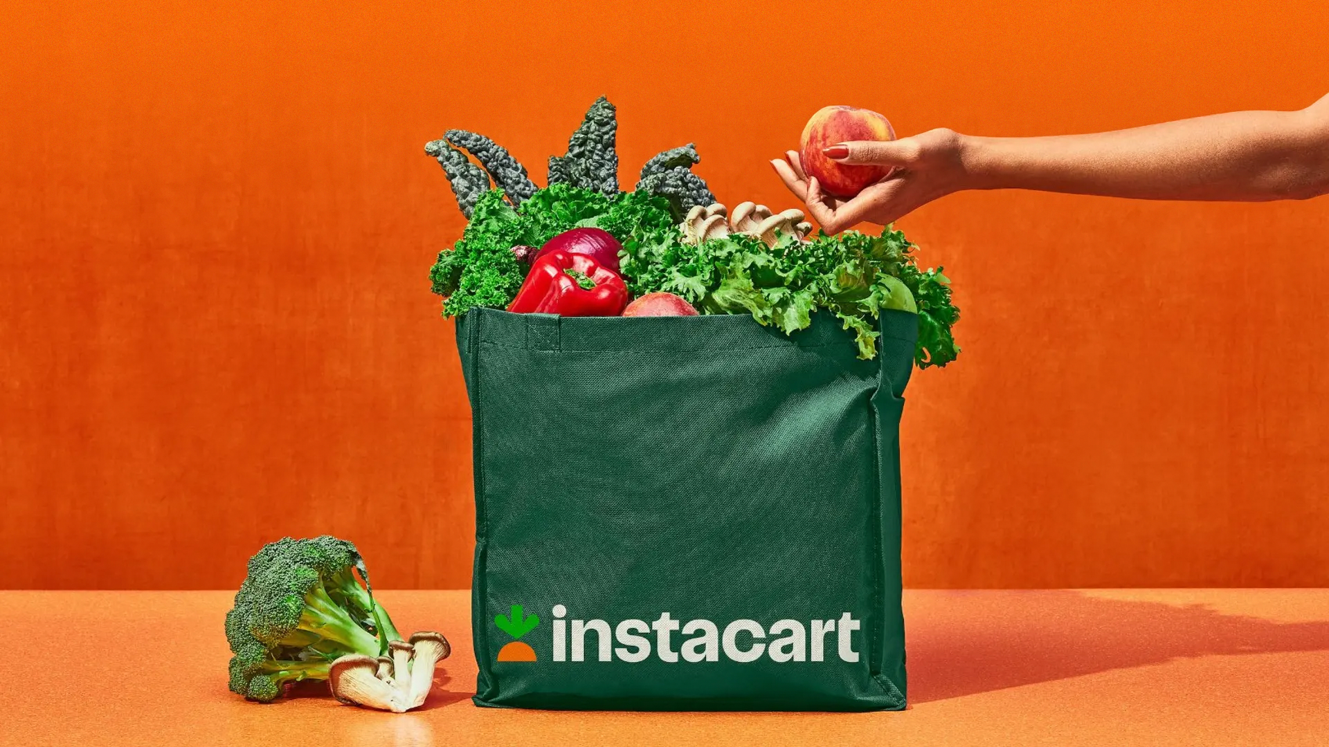 A hand adds an apple to a green Instacart bag full of fresh produce, with an orange backdrop.