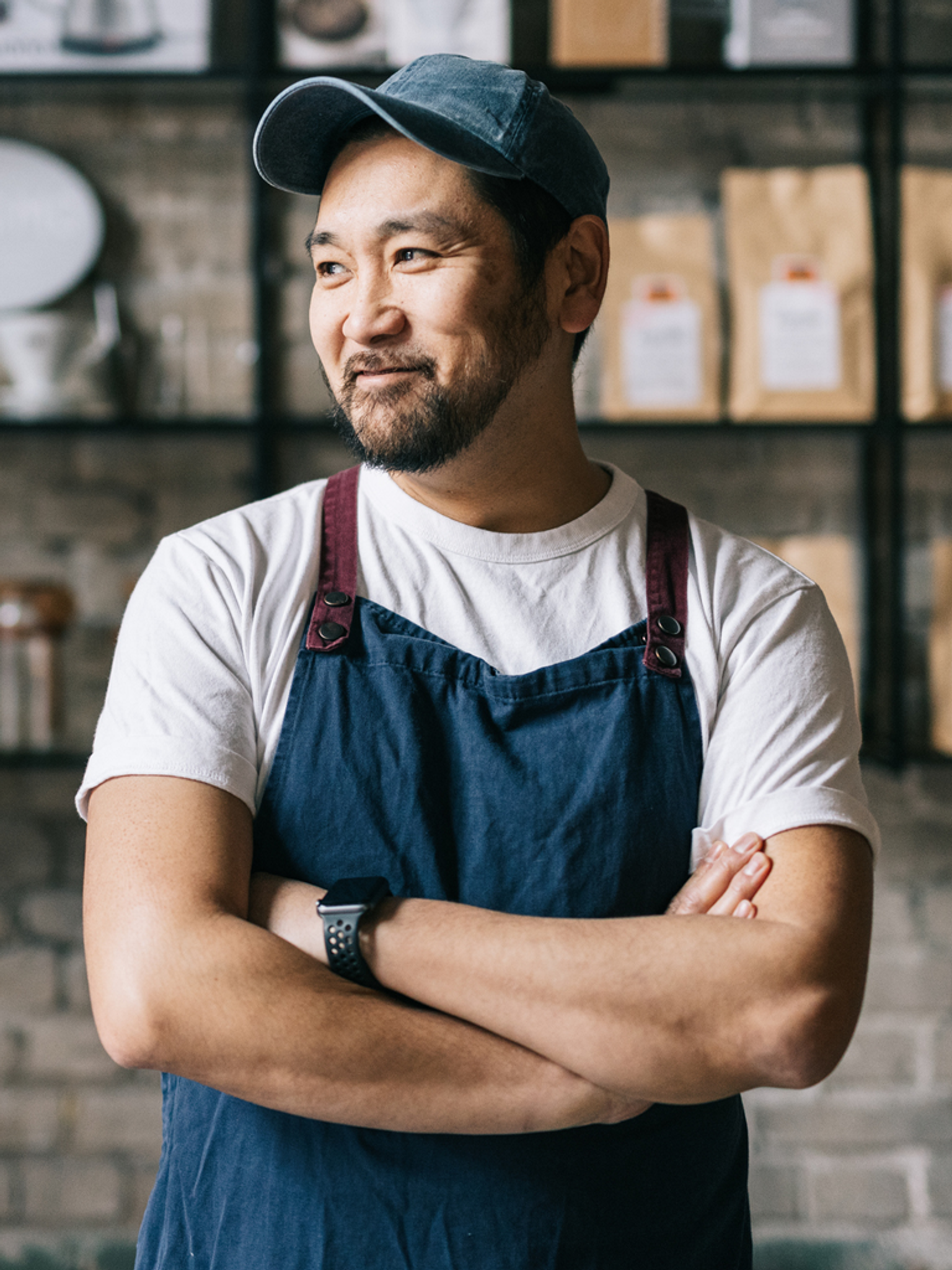 A man in an apron and cap, arms crossed, smiling slightly, stands in a shop with shelves of packaged goods behind him.