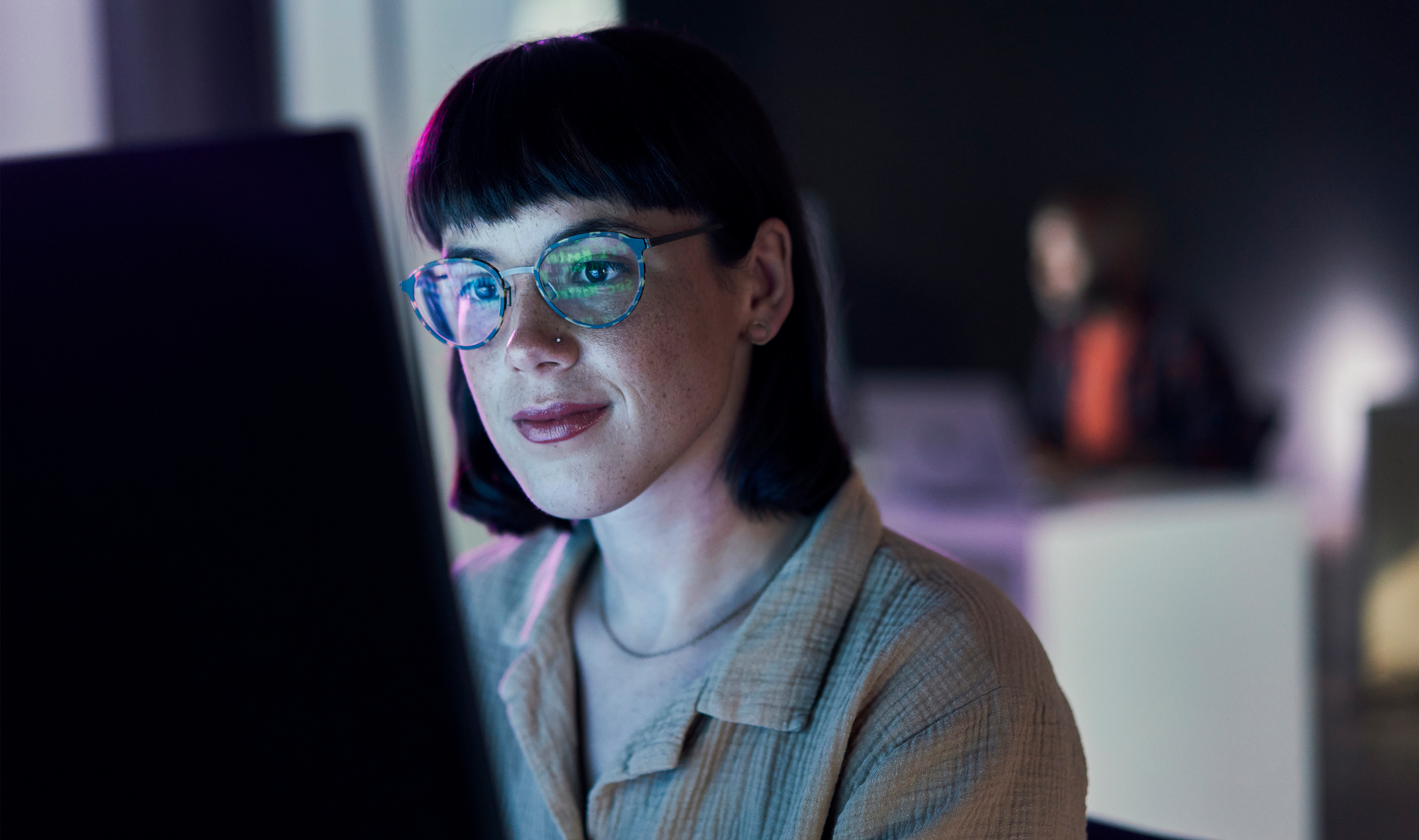 A woman with glasses smiles slightly while working at a computer in a blue-lit modern office.