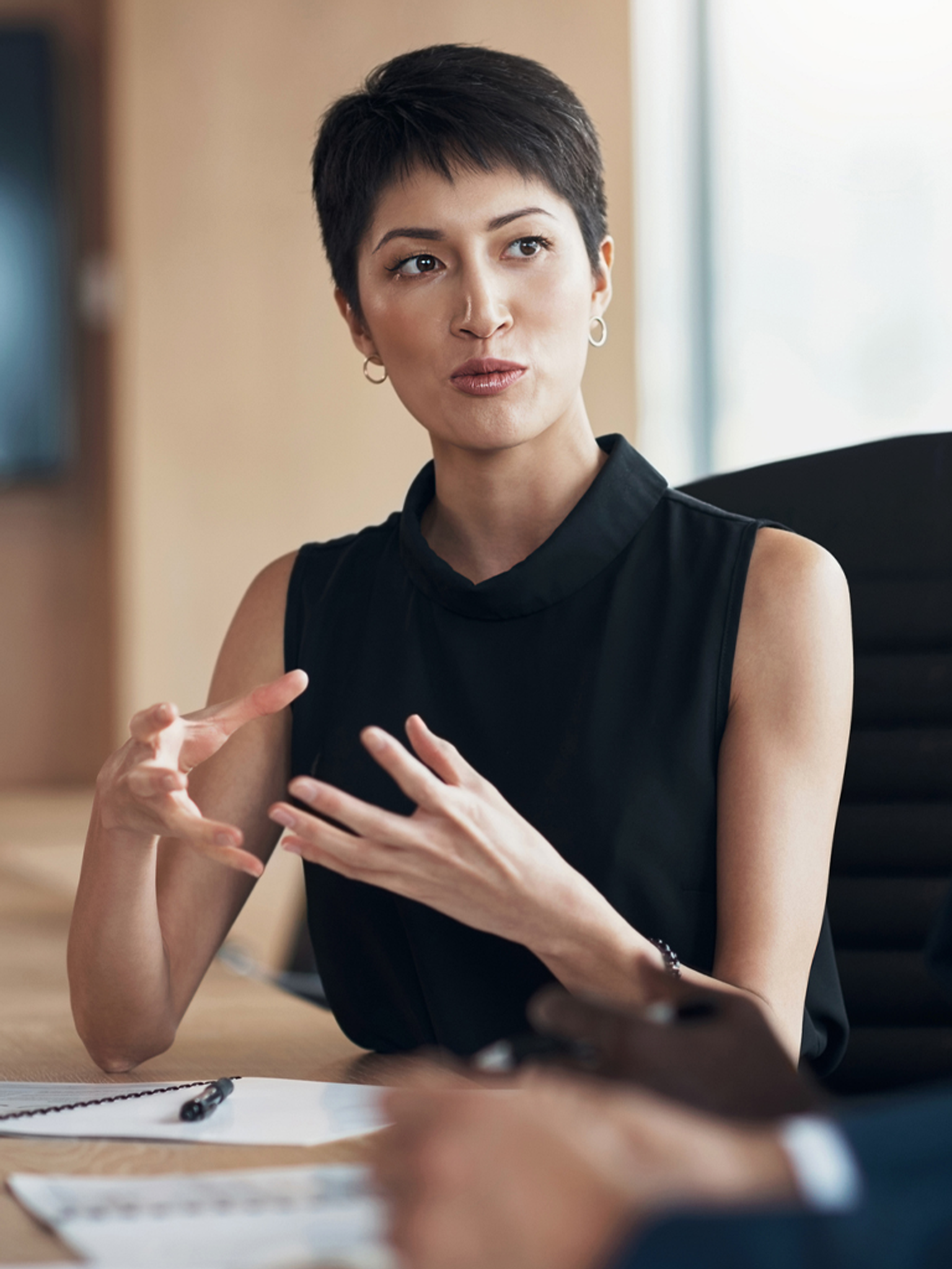 A professional woman with short hair gesticulating during a conversation, wearing a sleeveless top, with paperwork visible in front.