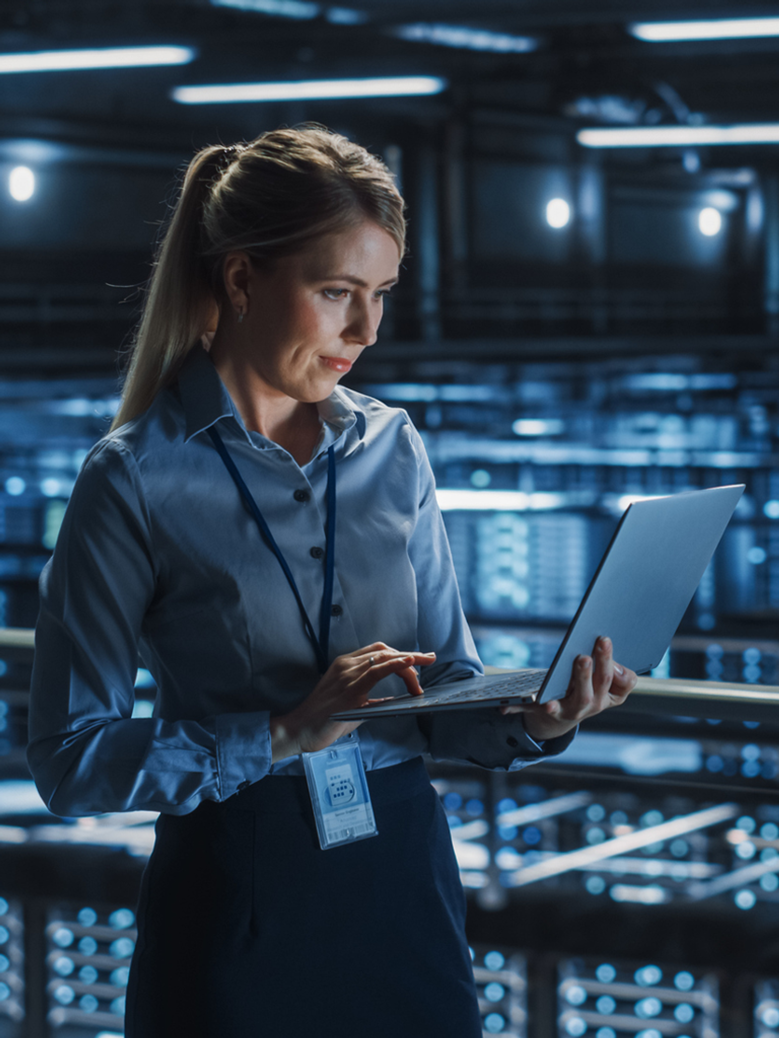 A professional woman with a laptop stands in a server room, focused on her work.