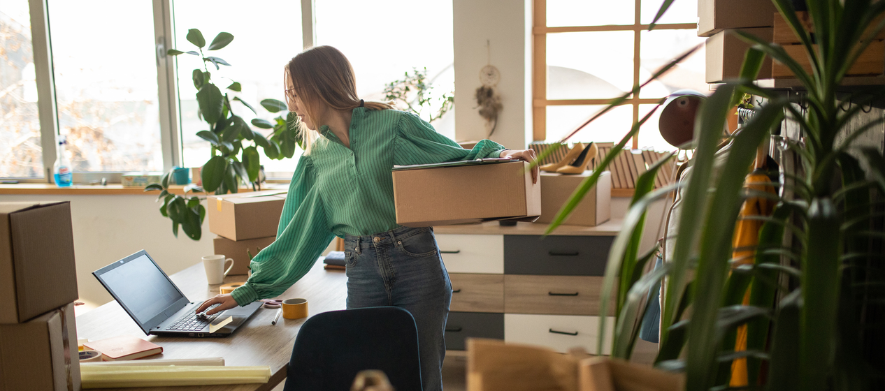 A person holding a box in an office with packing materials and a laptop on the desk.