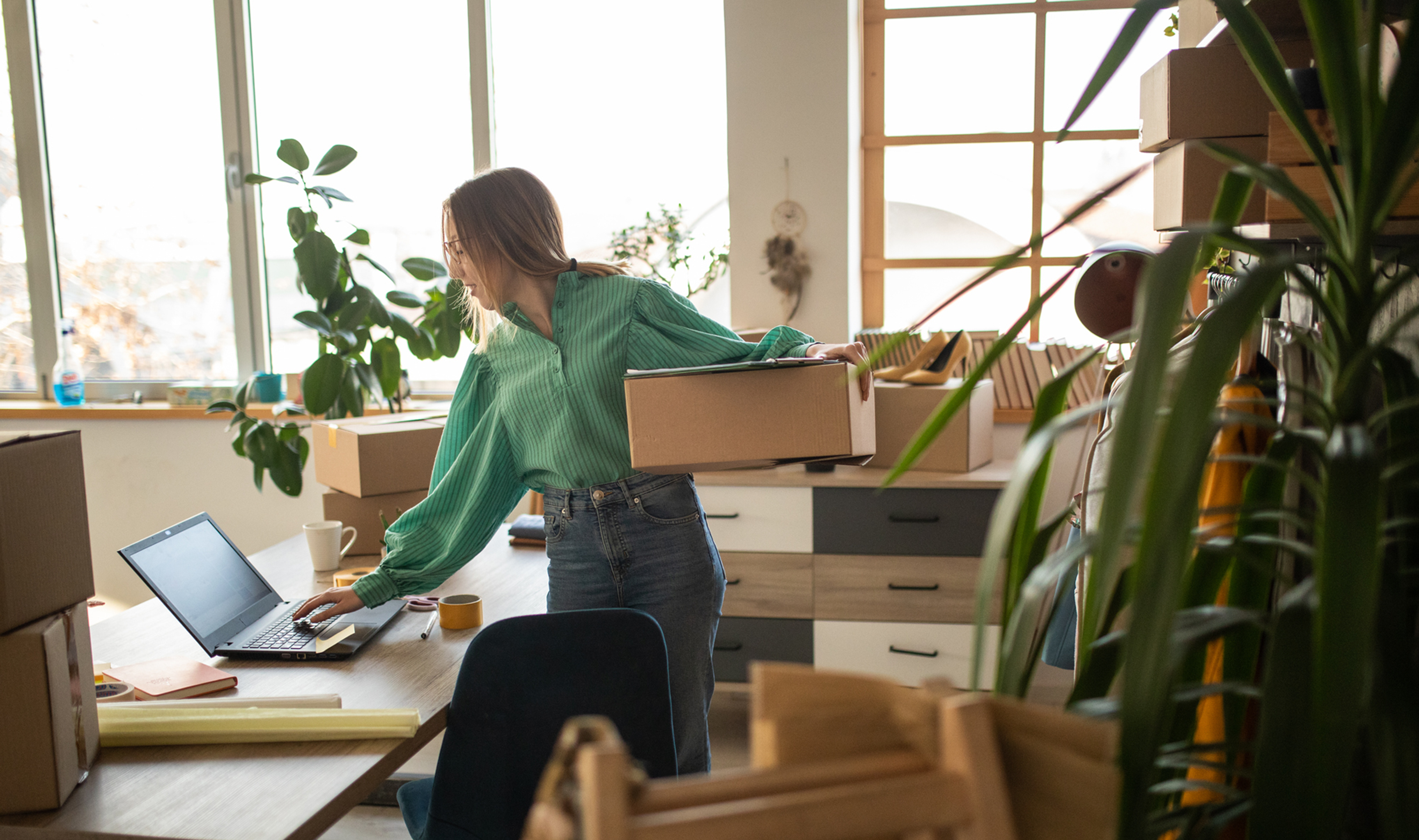 A person holding a box in an office with packing materials and a laptop on the desk.