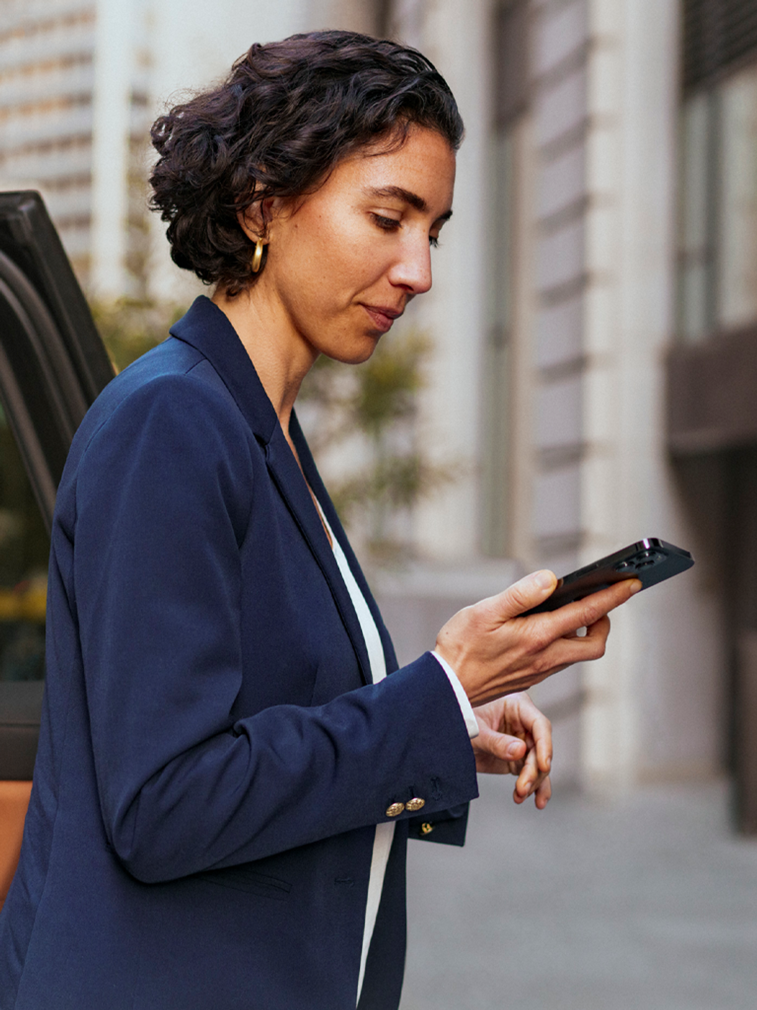 A person in a navy blazer looks at a smartphone intently on an urban street.