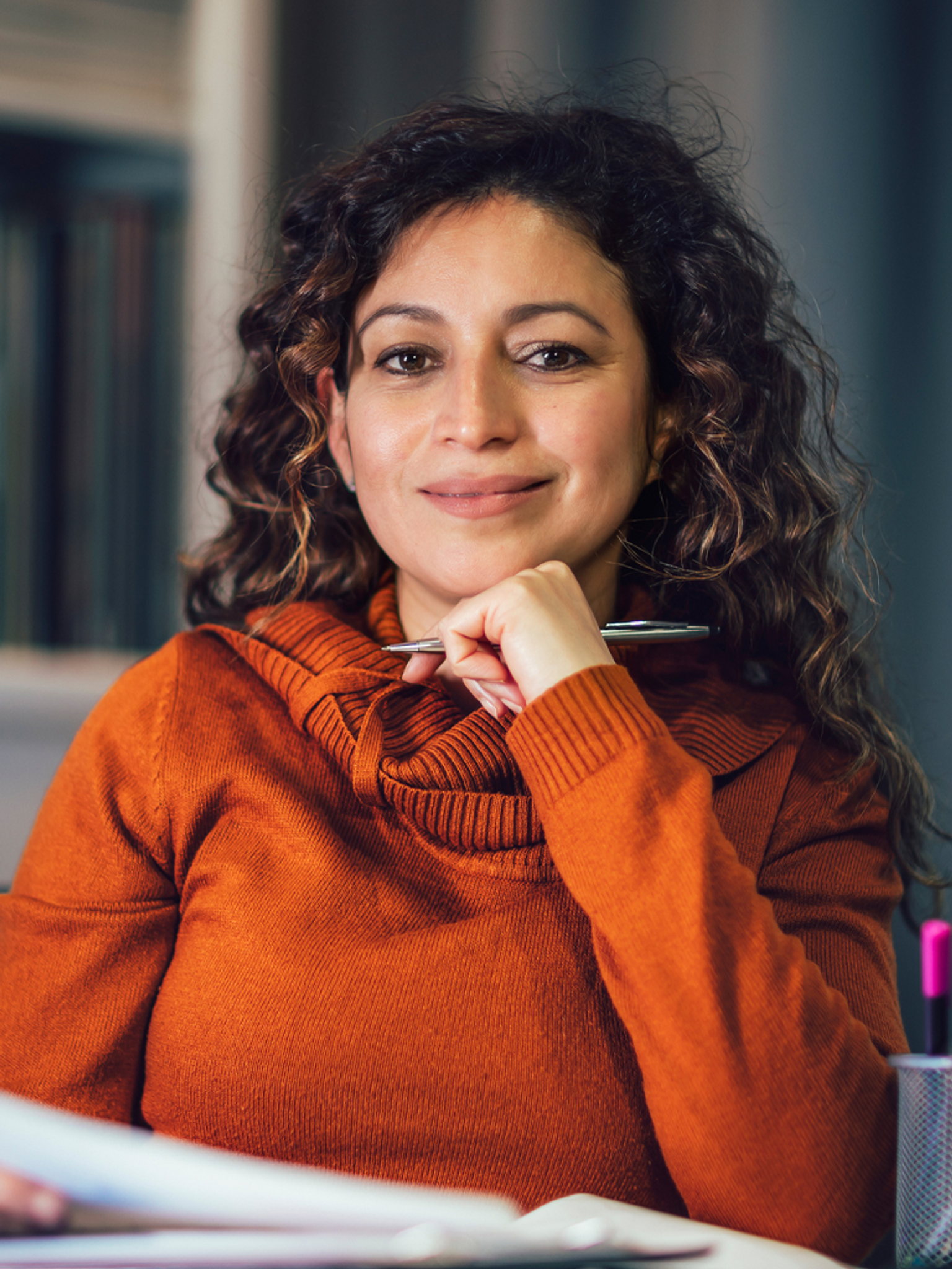 A smiling woman in an orange turtleneck holds a pen, resting her chin on her hand, with books nearby.